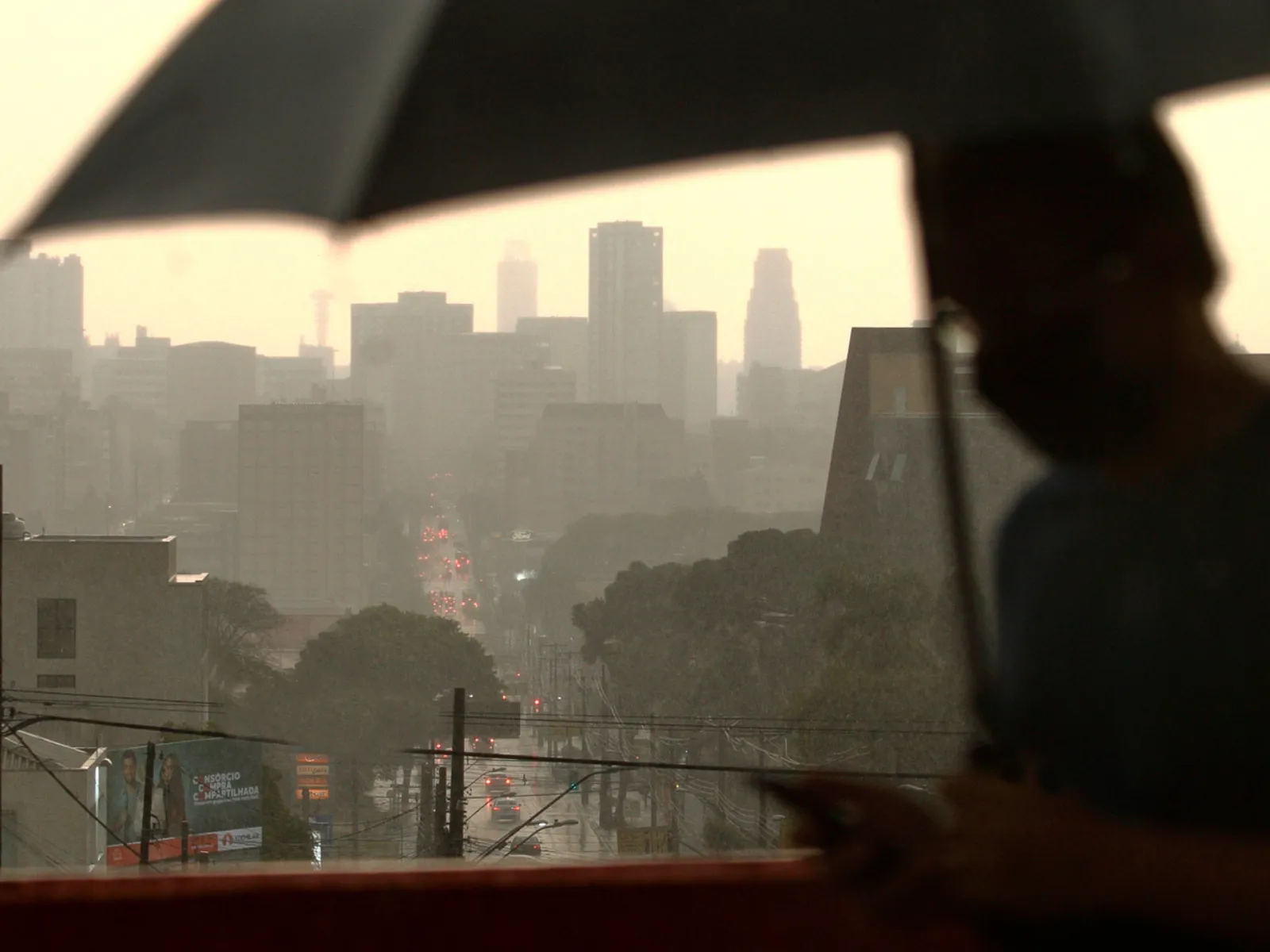 Fotografia de uma paisagem urbana vista sob chuva e névoa. Em primeiro plano, no lado direito e em silhueta desfocada, há uma pessoa segurando um guarda-chuva aberto e mexendo no celular. Ao fundo, vê-se uma avenida com trânsito de veículos e faróis acesos sob a chuva pesada, além do contorno de diversos edifícios altos envoltos por uma atmosfera acinzentada e névoa.