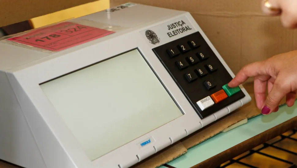 Fotografia em enquadramento fechado de uma urna eletrônica brasileira cinza sobre uma mesa. No lado direito do painel, uma mão com unhas pintadas de rosa acena o indicador pressionando a tecla verde "CONFIRMA". O teclado numérico preto traz também as teclas "BRANCO" e "CORRIGE". A tela retangular no lado esquerdo do aparelho está desligada (ou em branco).