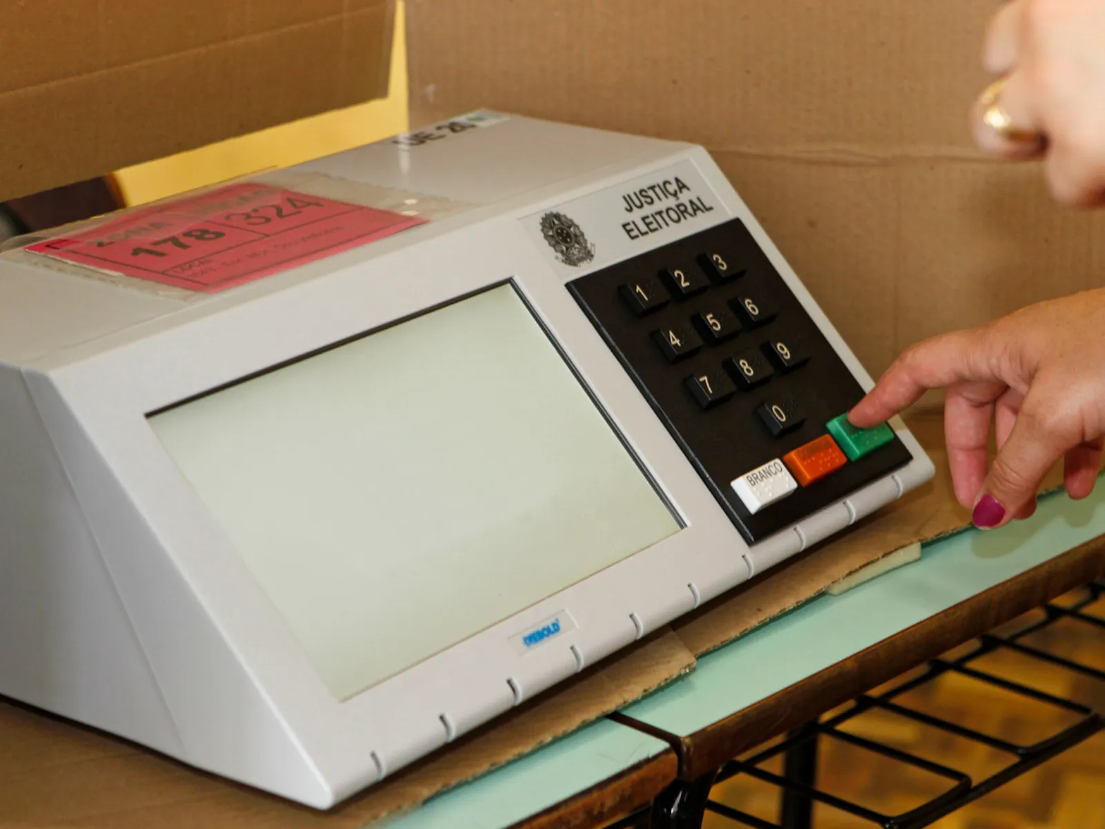 Fotografia em enquadramento fechado de uma urna eletrônica brasileira cinza sobre uma mesa. No lado direito do painel, uma mão com unhas pintadas de rosa acena o indicador pressionando a tecla verde "CONFIRMA". O teclado numérico preto traz também as teclas "BRANCO" e "CORRIGE". A tela retangular no lado esquerdo do aparelho está desligada (ou em branco).