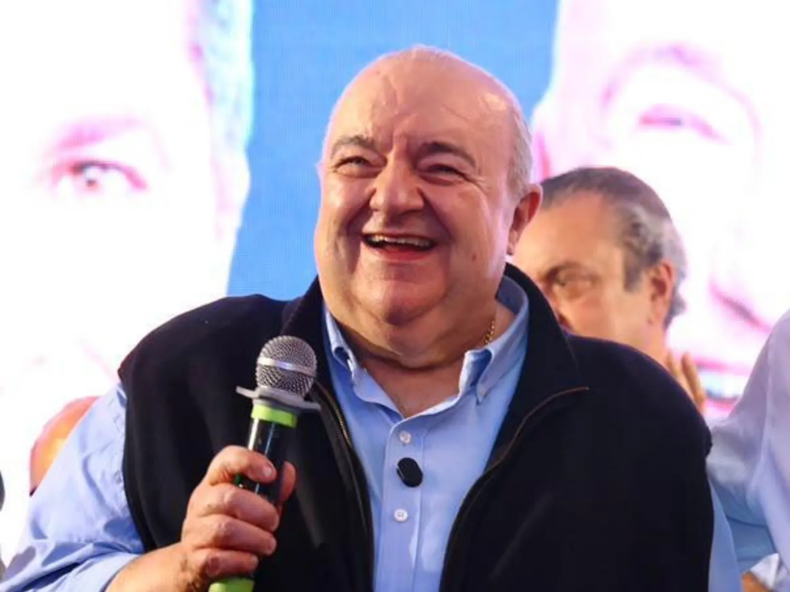 Fotografia em plano médio do ex-prefeito de Curitiba, Rafael Greca, um homem idoso de pele clara, calvo e com expressão sorridente. Ele segura um microfone com a mão direita perto do peito. Veste camisa social azul-clara e um colete escuro sobreposto. Ao fundo, ligeiramente desfocado, há um painel iluminado com imagens e outra pessoa ao seu lado.