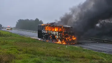 Fotografia de um ônibus escuro em chamas no acostamento de uma rodovia sob céu nublado. O veículo está tomado por fogo visível através das janelas e na parte dianteira, emitindo uma densa e grande coluna de fumaça preta que se espalha pelo ar. Em primeiro plano, há um canteiro de grama verde. Ao fundo, ao longo da estrada, veem-se uma ambulância e outros veículos parados com luzes de emergência ligadas, além de uma linha de árvores à beira da pista.