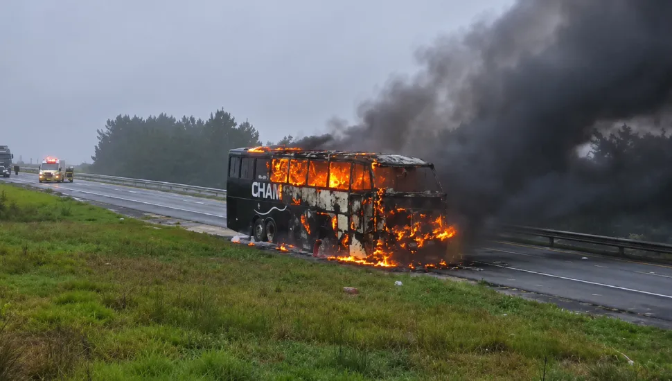 Fotografia de um ônibus escuro em chamas no acostamento de uma rodovia sob céu nublado. O veículo está tomado por fogo visível através das janelas e na parte dianteira, emitindo uma densa e grande coluna de fumaça preta que se espalha pelo ar. Em primeiro plano, há um canteiro de grama verde. Ao fundo, ao longo da estrada, veem-se uma ambulância e outros veículos parados com luzes de emergência ligadas, além de uma linha de árvores à beira da pista.