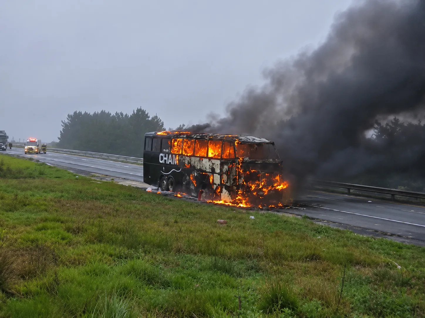Fotografia de um ônibus escuro em chamas no acostamento de uma rodovia sob céu nublado. O veículo está tomado por fogo visível através das janelas e na parte dianteira, emitindo uma densa e grande coluna de fumaça preta que se espalha pelo ar. Em primeiro plano, há um canteiro de grama verde. Ao fundo, ao longo da estrada, veem-se uma ambulância e outros veículos parados com luzes de emergência ligadas, além de uma linha de árvores à beira da pista.