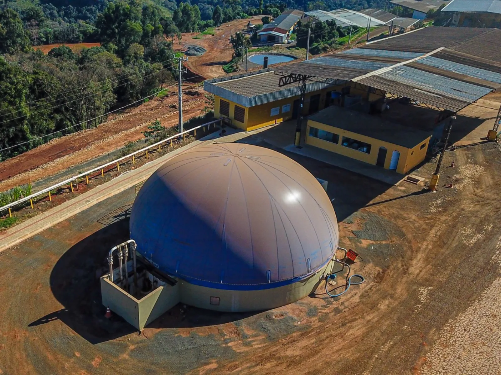 Fotografia em vista aérea de uma usina de biodigestão ou instalação industrial rural. Em primeiro plano, destaca-se um grande biodigestor circular com cúpula semiesférica em tom bege e base com faixa azul. Ao lado do tanque, há um suporte de concreto com tubulações metálicas. Ao fundo, veem-se galpões industriais com paredes amarelas e telhados metálicos, além de estradas de terra e vegetação em área rural ao redor.