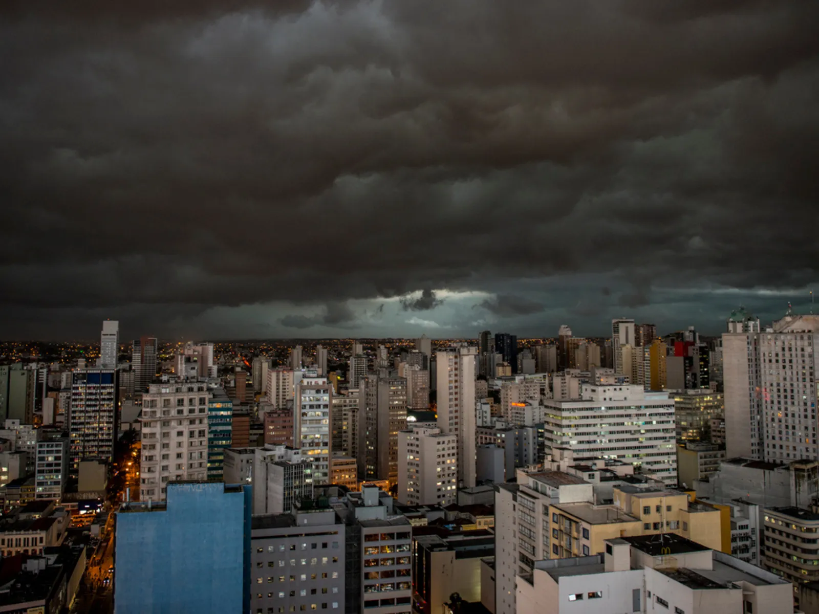 Fotografia em perspectiva elevada panorâmica de uma grande cidade sob tempestade. O céu está dominado por nuvens carregadas, escuras e densas em tons cinza-chumbo. Abaixo, estende-se um denso conjunto de edifícios urbanos com luzes acesas nas janelas e nas ruas, criando um contraste dramático entre o ambiente urbano iluminado e a atmosfera tempestuosa acima.