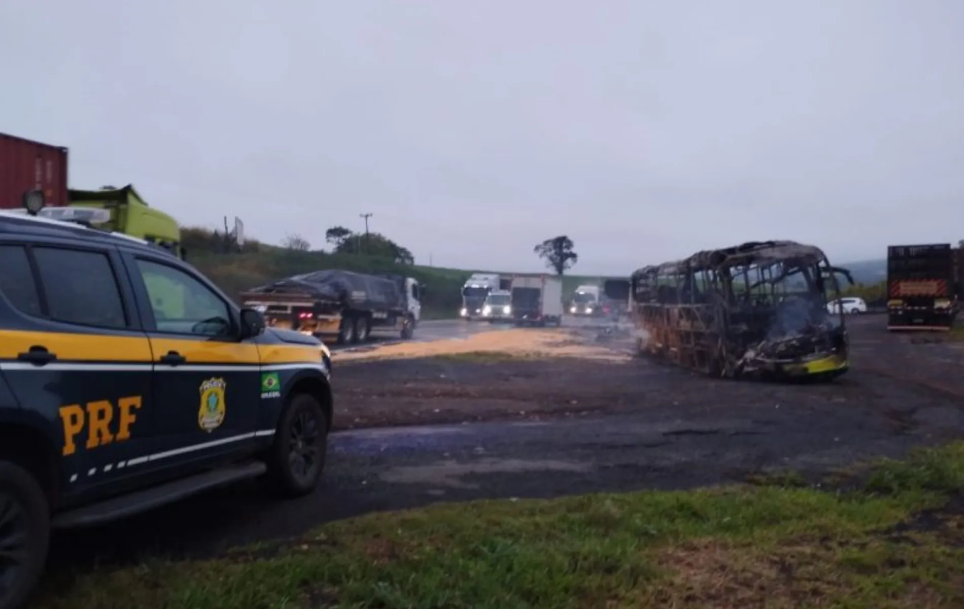Fotografia em campo aberto de um trecho de rodovia ao amanhecer sob céu nublado. Em primeiro plano, no lado esquerdo, vê-se a lateral de uma viatura da Polícia Rodoviária Federal (PRF), preta e amarela, com o brasão e a sigla em destaque. À direita, no acostamento, está a carcaça completamente queimada e destruída de um ônibus. Ao fundo, o trânsito de caminhões e outros veículos segue retido pela pista sob luzes acesas.