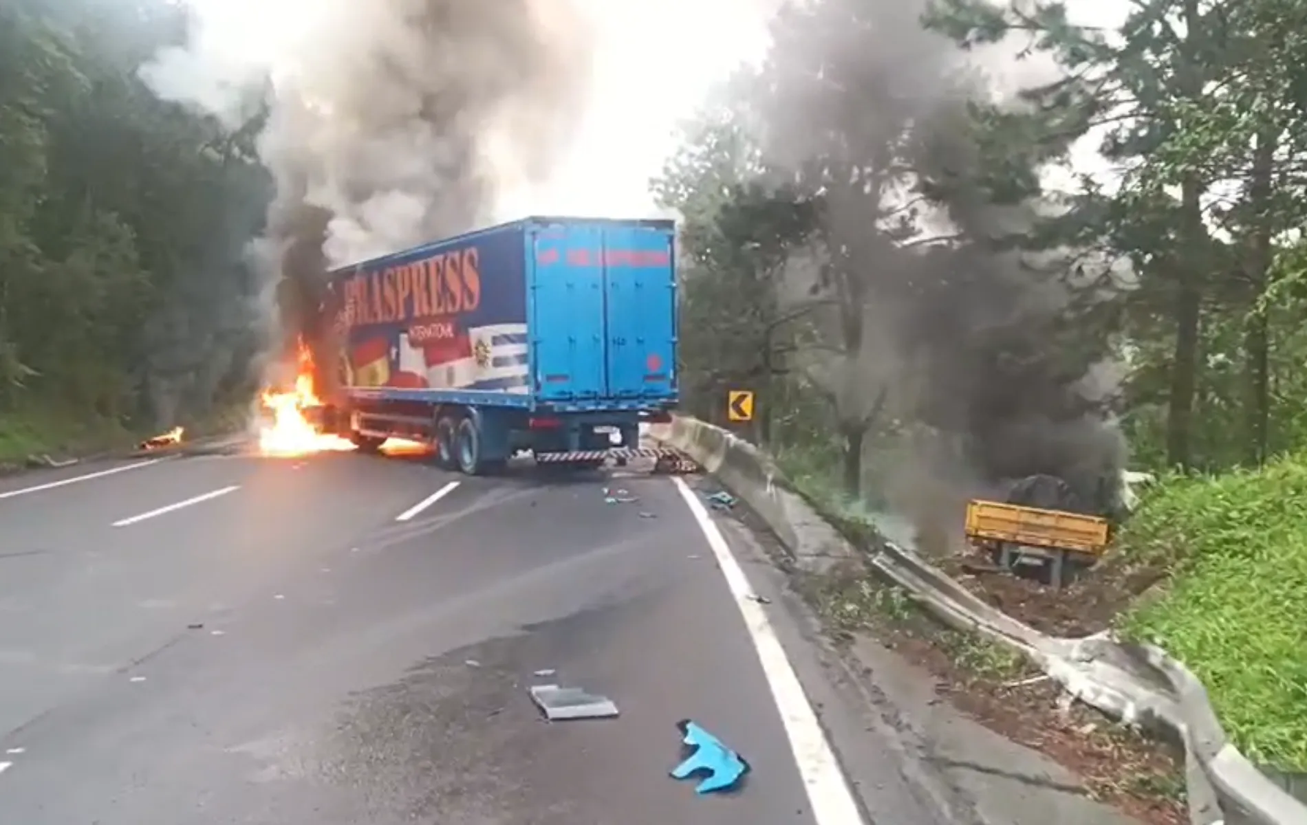 Fotografia de um acidente de trânsito em uma rodovia sinuosa cercada por vegetação densa. No centro da pista, um caminhão baú azul da empresa Braspress está parado, com a cabine tomada por fogo visível e chamas altas, gerando uma grande coluna de fumaça cinza. À direita, fora da pista e caído em um barranco atrás da mureta de proteção, há outro veículo envolvido no acidente. Destroços e peças plásticas azuis estão espalhados pelo asfalto.