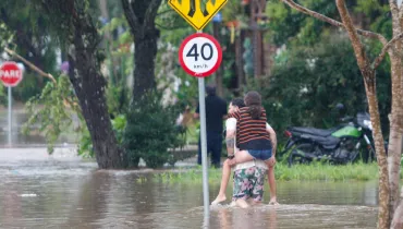 Fotografia em plano aberto mostrando uma rua alagada por água barrenta durante ou após uma tempestade. No centro da imagem, um homem caminha com a água acima dos tornozelos carregando uma jovem nas costas para atravessar o trecho inundado. A jovem veste uma blusa listrada e shorts jeans. Próximo a eles, há uma placa circular de trânsito indicando limite de velocidade de "40 km/h". Ao fundo, veem-se árvores, uma placa de "PARE" e uma motocicleta estacionada na calçada.