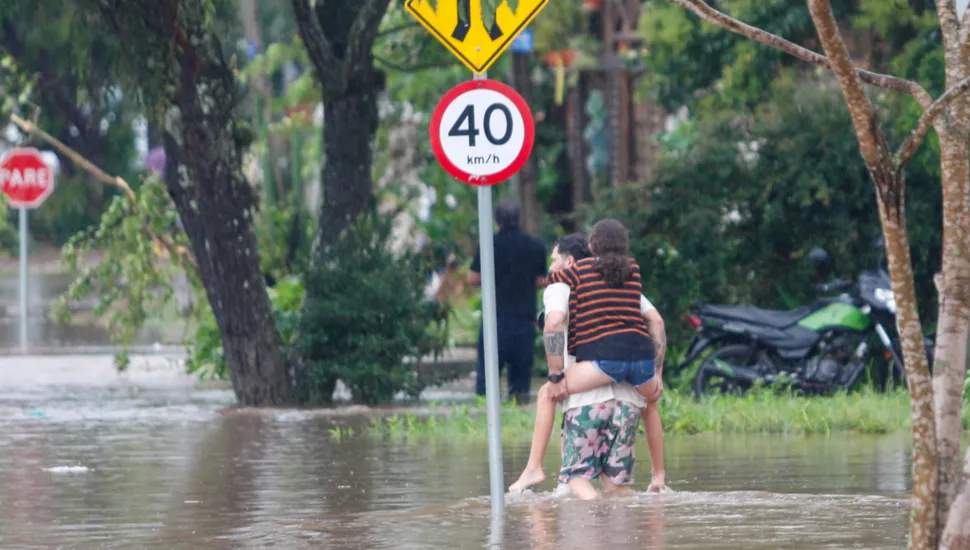 Fotografia em plano aberto mostrando uma rua alagada por água barrenta durante ou após uma tempestade. No centro da imagem, um homem caminha com a água acima dos tornozelos carregando uma jovem nas costas para atravessar o trecho inundado. A jovem veste uma blusa listrada e shorts jeans. Próximo a eles, há uma placa circular de trânsito indicando limite de velocidade de "40 km/h". Ao fundo, veem-se árvores, uma placa de "PARE" e uma motocicleta estacionada na calçada.