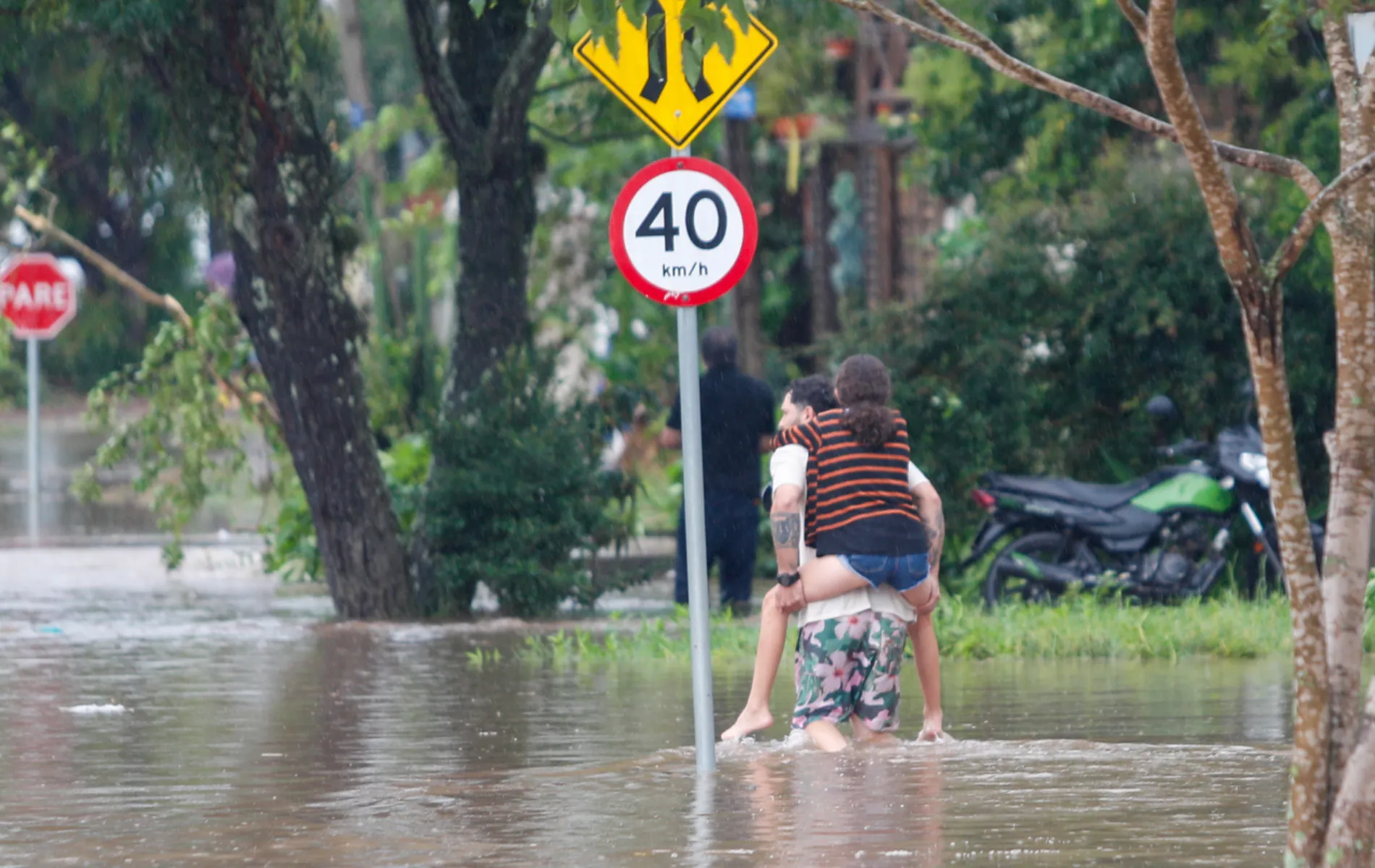 Fotografia em plano aberto mostrando uma rua alagada por água barrenta durante ou após uma tempestade. No centro da imagem, um homem caminha com a água acima dos tornozelos carregando uma jovem nas costas para atravessar o trecho inundado. A jovem veste uma blusa listrada e shorts jeans. Próximo a eles, há uma placa circular de trânsito indicando limite de velocidade de "40 km/h". Ao fundo, veem-se árvores, uma placa de "PARE" e uma motocicleta estacionada na calçada.