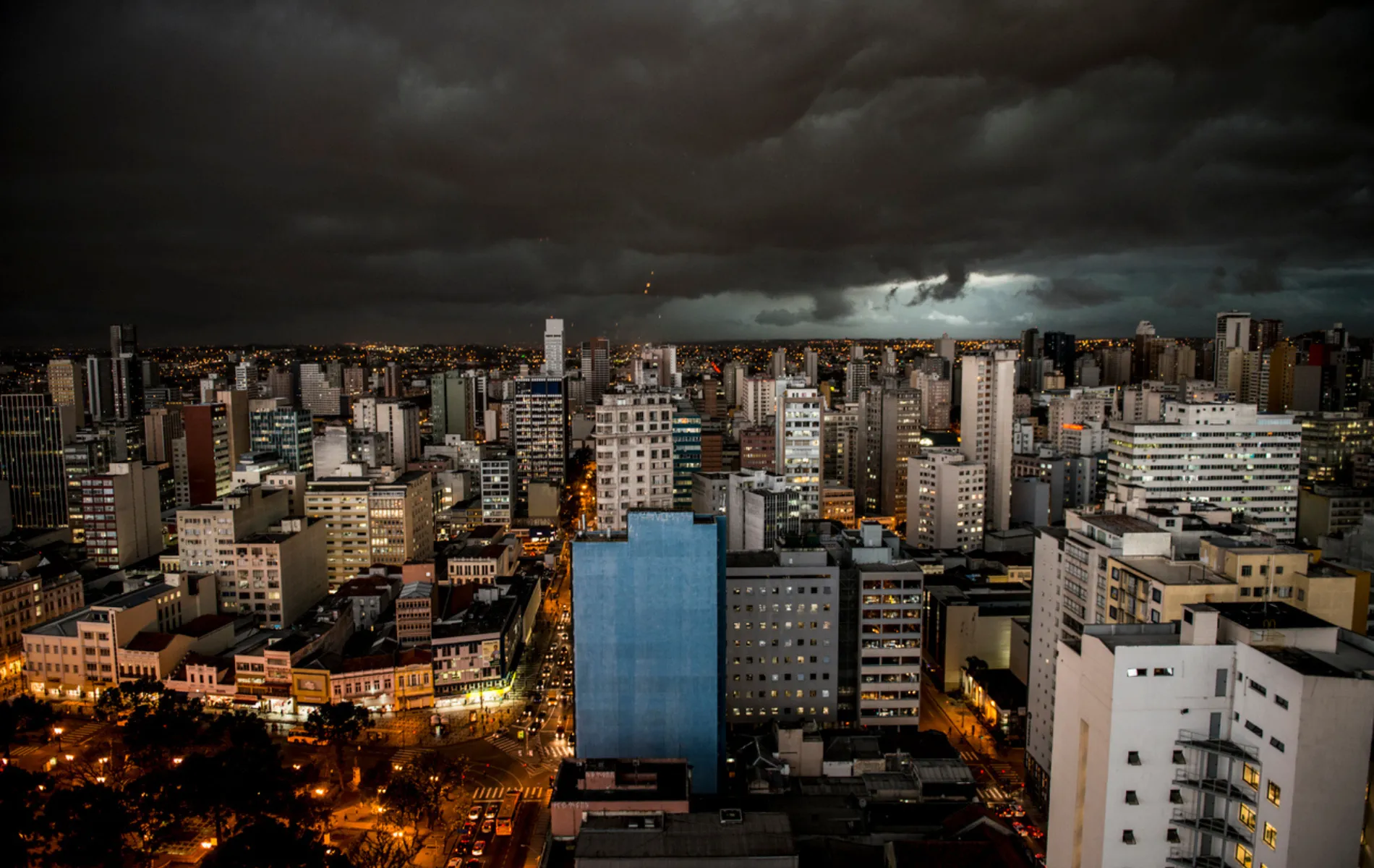 Fotografia panorâmica em vista aérea noturna de um denso centro urbano sob tempestade. O céu na metade superior está coberto por nuvens escuras e tempestuosas em tons de cinza-chumbo e preto. Abaixo, a paisagem urbana exibe múltiplos edifícios e prédios residenciais e comerciais com janelas iluminadas. No canto inferior esquerdo e ao longo das avenidas, as luzes amareladas e alaranjadas dos postes e dos veículos criam um contraste vibrante com o céu carregado.