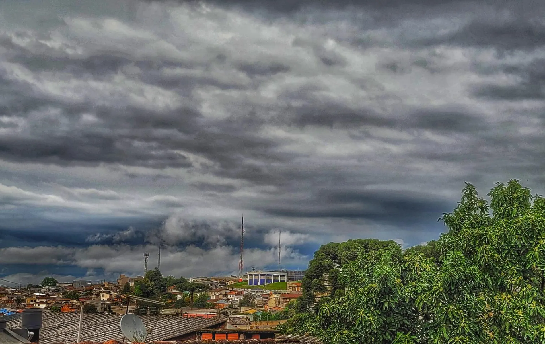 Fotografia em perspectiva ampla mostrando o céu de um bairro residencial sob nuvens de tempestade. O céu está coberto por formações de nuvens densas, onduladas e acinzentadas. Em primeiro plano, no canto inferior direito, destacam-se as copas de árvores verdes e exuberantes. No canto inferior esquerdo, veem-se os telhados de casas residenciais com uma antena parabólica. Ao fundo, no horizonte sob a tempestade, estende-se o bairro com antenas de transmissão e construções urbanas.