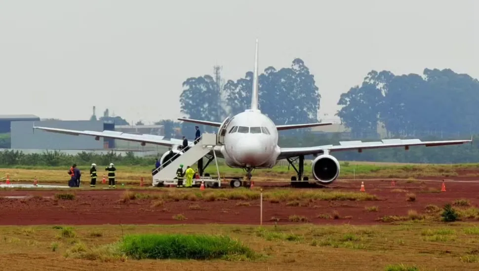 Fotografia frontal de um avião de passageiros de grande porte, predominantemente branco, parado fora da pista de pouso em uma área de solo de terra vermelha e gramado de um aeroporto. A aeronave apresenta uma leve inclinação lateral. Uma escada móvel de desembarque está acoplada à porta dianteira esquerda, por onde pessoas descem. Ao redor da aeronave e na base da escada, veem-se bombeiros e equipes de emergência uniformizadas em atendimento. Ao fundo, sob céu claro e nublado, há uma linha de árvores e instalações aeroportuárias.