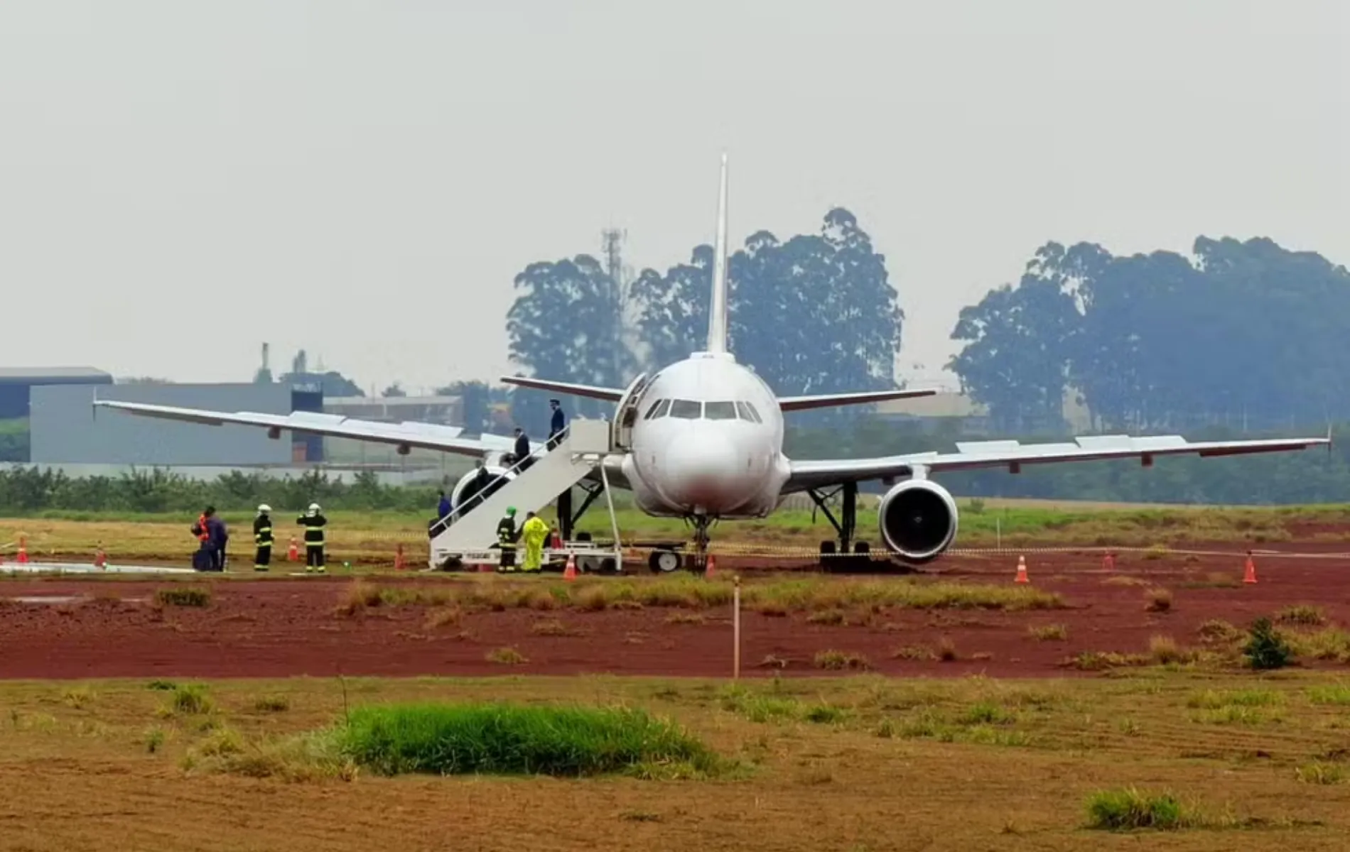 Fotografia frontal de um avião de passageiros de grande porte, predominantemente branco, parado fora da pista de pouso em uma área de solo de terra vermelha e gramado de um aeroporto. A aeronave apresenta uma leve inclinação lateral. Uma escada móvel de desembarque está acoplada à porta dianteira esquerda, por onde pessoas descem. Ao redor da aeronave e na base da escada, veem-se bombeiros e equipes de emergência uniformizadas em atendimento. Ao fundo, sob céu claro e nublado, há uma linha de árvores e instalações aeroportuárias.