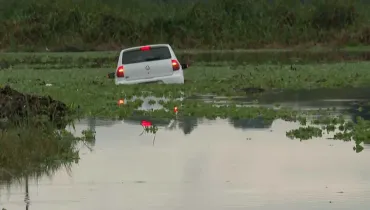 Fotografia em enquadramento aberto mostrando um carro hatch branco, da marca Volkswagen, parcialmente submerso em um corpo d'água coberto por vegetação aquática e aguapés. Visto de traseira, o veículo está com as luzes dos faróis e lanternas acesas, refletindo na superfície da água. Ao fundo e nas laterais, estende-se uma área com vegetação rasteira e alta sob iluminação natural suave.