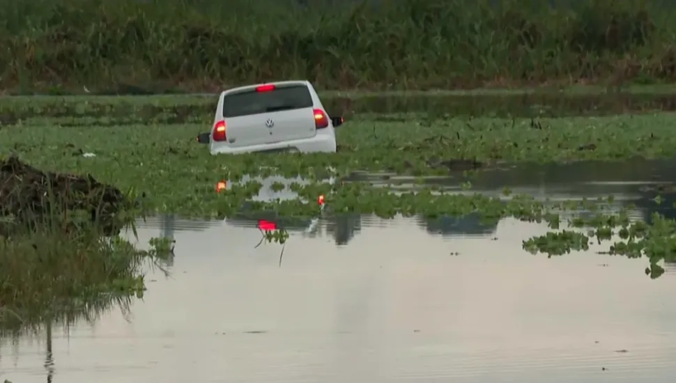 #PraCegoVer #DescriçãoDaImagem Fotografia em enquadramento aberto mostrando um carro hatch branco, da marca Volkswagen, parcialmente submerso em um corpo d'água coberto por vegetação aquática e aguapés. Visto de traseira, o veículo está com as luzes dos faróis e lanternas acesas, refletindo na superfície da água. Ao fundo e nas laterais, estende-se uma área com vegetação rasteira e alta sob iluminação natural suave.