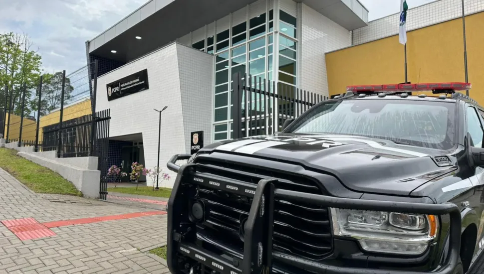 Fotografia em ângulo diagonal focando a dianteira de uma caminhonete preta da Polícia Civil do Paraná (PCPR), equipada com para-choque de impulsão metálico e giroflex vermelho no teto. O veículo está estacionado em frente ao prédio moderno de uma delegacia da PCPR, que possui fachada de pastilhas brancas, janelas espelhadas e um painel preto com o logotipo e a identificação da instituição. No lado esquerdo, vê-se uma calçada com piso tátil vermelho sob céu ligeiramente nublado.