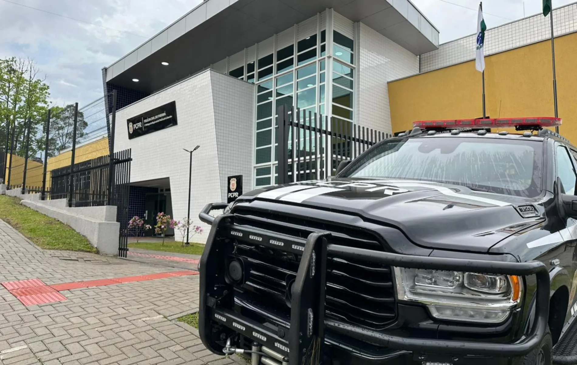 Fotografia em ângulo diagonal focando a dianteira de uma caminhonete preta da Polícia Civil do Paraná (PCPR), equipada com para-choque de impulsão metálico e giroflex vermelho no teto. O veículo está estacionado em frente ao prédio moderno de uma delegacia da PCPR, que possui fachada de pastilhas brancas, janelas espelhadas e um painel preto com o logotipo e a identificação da instituição. No lado esquerdo, vê-se uma calçada com piso tátil vermelho sob céu ligeiramente nublado.
