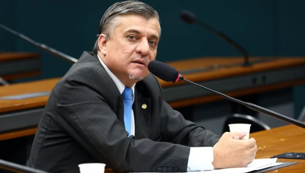 Fotografia em plano médio do deputado federal José Guimarães, um homem de pele clara com cabelos escuros penteados para trás e cavanhaque grisalho. Ele está sentado à mesa de um plenário ou comissão governamental, levemente inclinado para a frente e falando ao microfone com haste flexível. Veste terno risca-de-giz escuro, camisa branca, gravata azul-clara e um broche oficial na lapela. À sua frente, sobre a mesa de madeira, há papéis, uma caneta azul e dois copos plásticos descartáveis brancos.