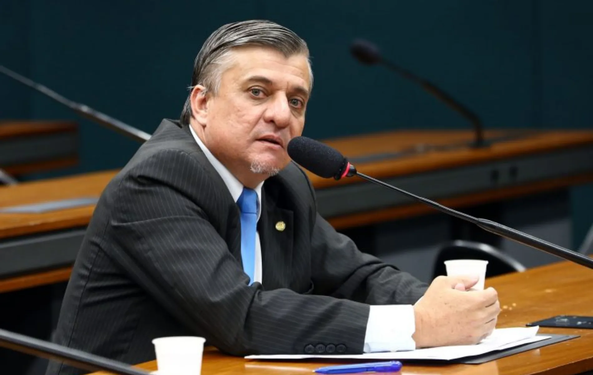 Fotografia em plano médio do deputado federal José Guimarães, um homem de pele clara com cabelos escuros penteados para trás e cavanhaque grisalho. Ele está sentado à mesa de um plenário ou comissão governamental, levemente inclinado para a frente e falando ao microfone com haste flexível. Veste terno risca-de-giz escuro, camisa branca, gravata azul-clara e um broche oficial na lapela. À sua frente, sobre a mesa de madeira, há papéis, uma caneta azul e dois copos plásticos descartáveis brancos.