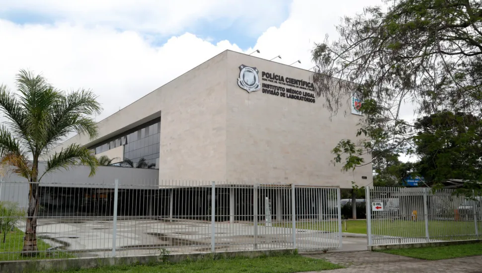 Fotografia em plano aberto do prédio da Polícia Científica e Instituto Médico Legal (IML) do Paraná. A edificação moderna possui fachada revestida em blocos bege e janelas de vidro espelhado escuro. No topo da fachada, lê-se em letras escuras a identificação "POLÍCIA CIENTÍFICA - INSTITUTO MÉDICO LEGAL - DIVISÃO DE LABORATÓRIOS" acompanhada pelo brasão oficial. O edifício é cercado por um gradil metálico cinza e portão de acesso. Em primeiro plano, à esquerda, há uma pequena palmeira e trecho de calçada sob céu parcialmente nublado.