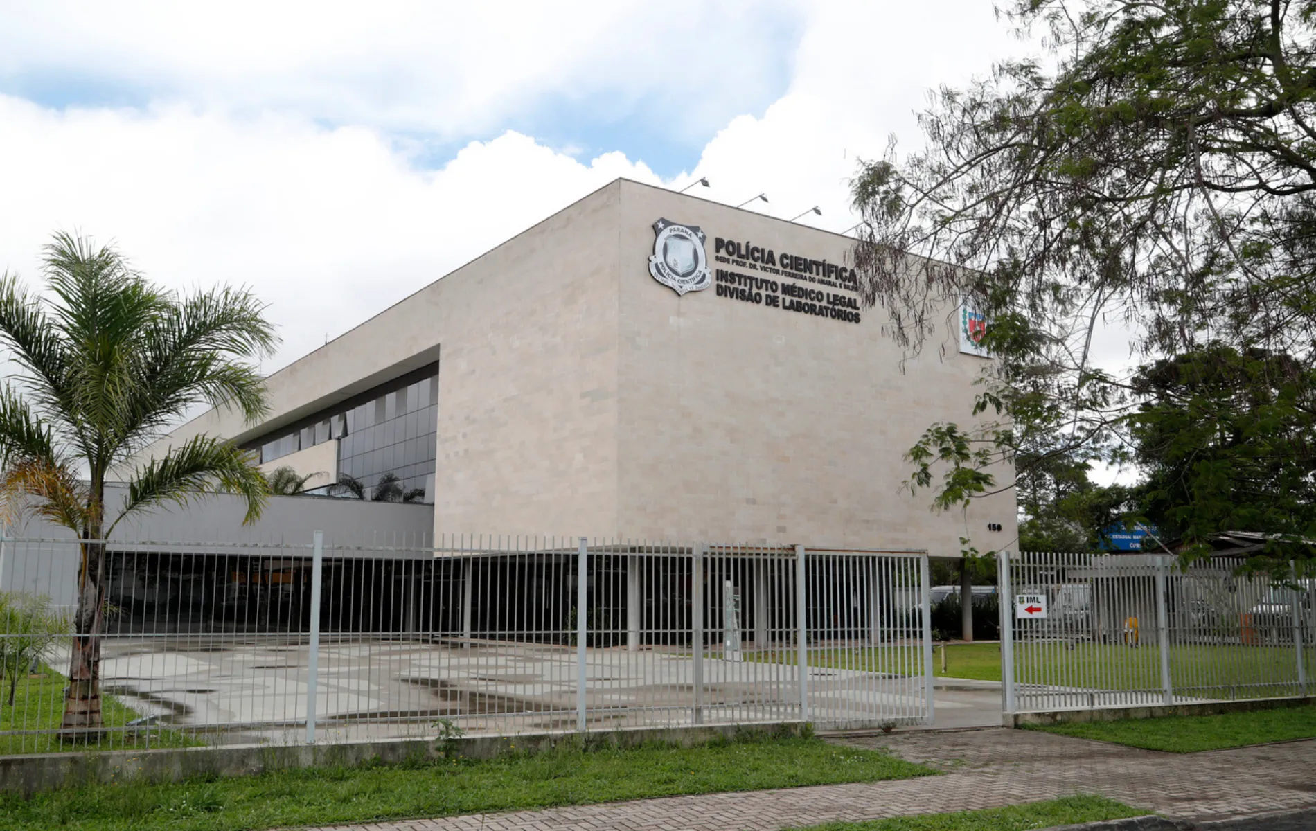Fotografia em plano aberto do prédio da Polícia Científica e Instituto Médico Legal (IML) do Paraná. A edificação moderna possui fachada revestida em blocos bege e janelas de vidro espelhado escuro. No topo da fachada, lê-se em letras escuras a identificação "POLÍCIA CIENTÍFICA - INSTITUTO MÉDICO LEGAL - DIVISÃO DE LABORATÓRIOS" acompanhada pelo brasão oficial. O edifício é cercado por um gradil metálico cinza e portão de acesso. Em primeiro plano, à esquerda, há uma pequena palmeira e trecho de calçada sob céu parcialmente nublado.