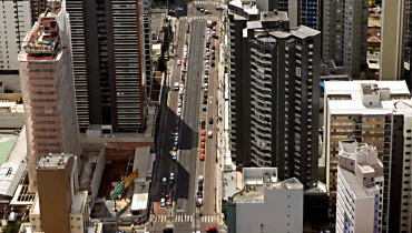 Fotografia em vista aérea superior (vista de cima) de uma grande avenida urbana ladeada por diversos edifícios e arranha-céus. A avenida corta o centro do enquadramento na vertical, com carros em movimento e estacionados ao longo das faixas e calçadas. À esquerda, destaca-se um edifício em construção coberto por rede de proteção clara. Ao redor, há prédios residenciais e comerciais de diferentes alturas, cores e estilos arquitetônicos sob luz solar direta, projetando sombras marcadas sobre o asfalto.