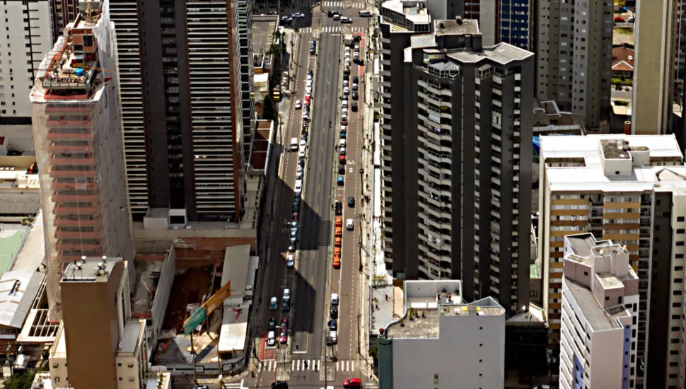 Fotografia em vista aérea superior (vista de cima) de uma grande avenida urbana ladeada por diversos edifícios e arranha-céus. A avenida corta o centro do enquadramento na vertical, com carros em movimento e estacionados ao longo das faixas e calçadas. À esquerda, destaca-se um edifício em construção coberto por rede de proteção clara. Ao redor, há prédios residenciais e comerciais de diferentes alturas, cores e estilos arquitetônicos sob luz solar direta, projetando sombras marcadas sobre o asfalto.