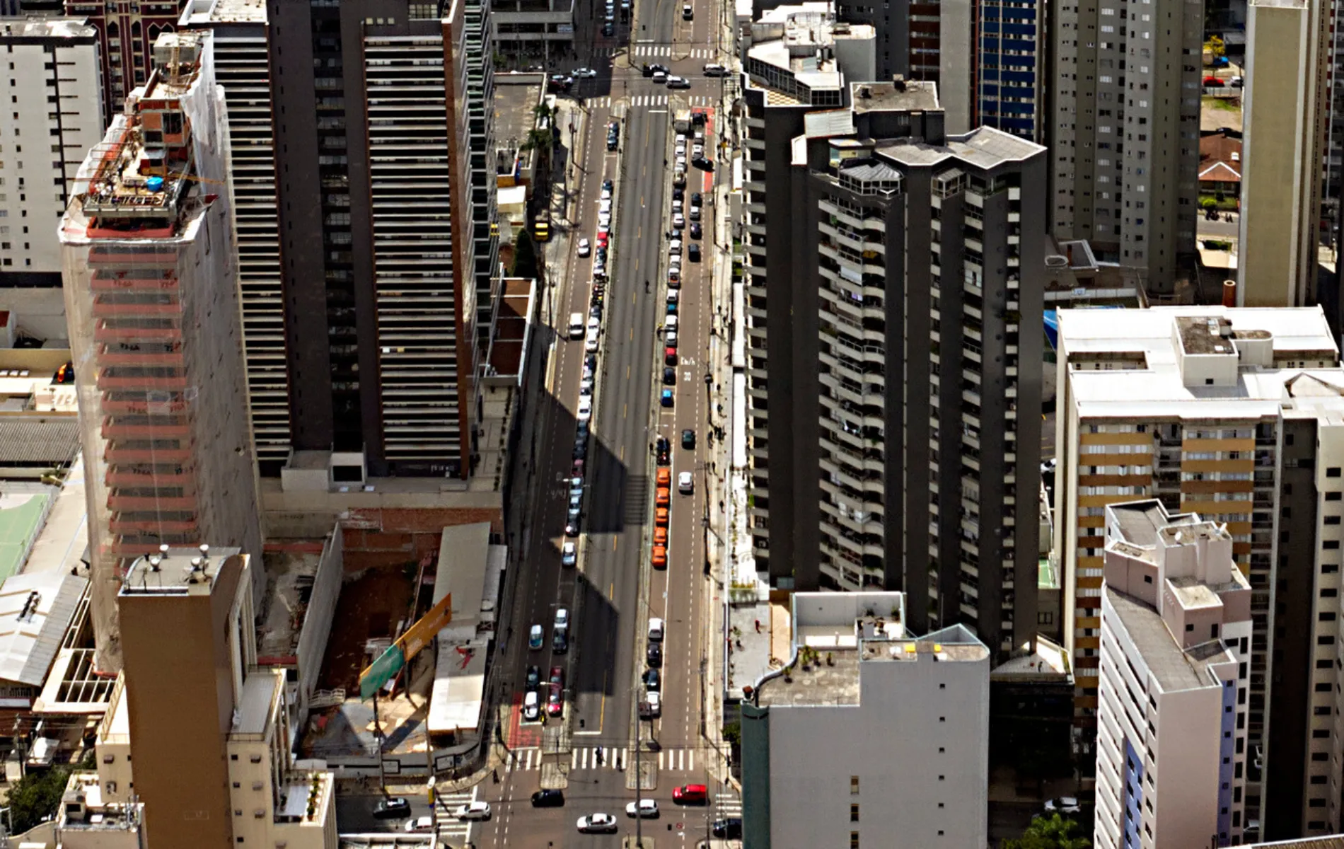 Fotografia em vista aérea superior (vista de cima) de uma grande avenida urbana ladeada por diversos edifícios e arranha-céus. A avenida corta o centro do enquadramento na vertical, com carros em movimento e estacionados ao longo das faixas e calçadas. À esquerda, destaca-se um edifício em construção coberto por rede de proteção clara. Ao redor, há prédios residenciais e comerciais de diferentes alturas, cores e estilos arquitetônicos sob luz solar direta, projetando sombras marcadas sobre o asfalto.