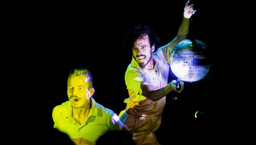 Fotografia de cena teatral com fundo inteiramente preto. Em primeiro plano, dois homens de pele clara estão em movimento sob iluminação cênica vibrante em tons amarelados e azulados. À direita, um homem de cabelos cacheados e barba sorri expressivamente enquanto segura um globo de espelhos prateado elevado com uma das mãos. À esquerda, em plano levemente rebaixado, outro homem de cabelo curto e bigode aparece com expressão de surpresa e boca aberta, vestindo camisa de gola polo amarela.