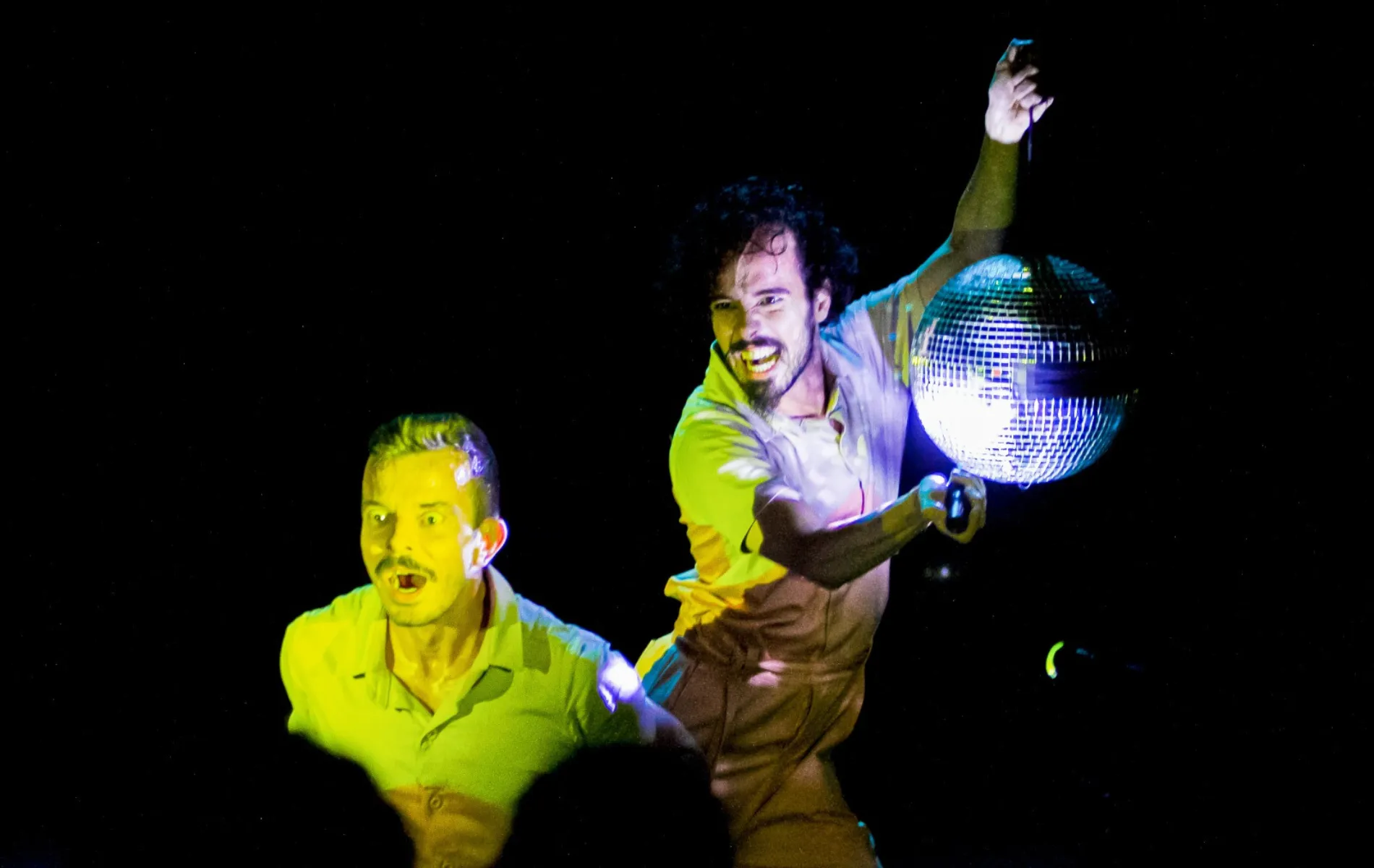 Fotografia de cena teatral com fundo inteiramente preto. Em primeiro plano, dois homens de pele clara estão em movimento sob iluminação cênica vibrante em tons amarelados e azulados. À direita, um homem de cabelos cacheados e barba sorri expressivamente enquanto segura um globo de espelhos prateado elevado com uma das mãos. À esquerda, em plano levemente rebaixado, outro homem de cabelo curto e bigode aparece com expressão de surpresa e boca aberta, vestindo camisa de gola polo amarela.