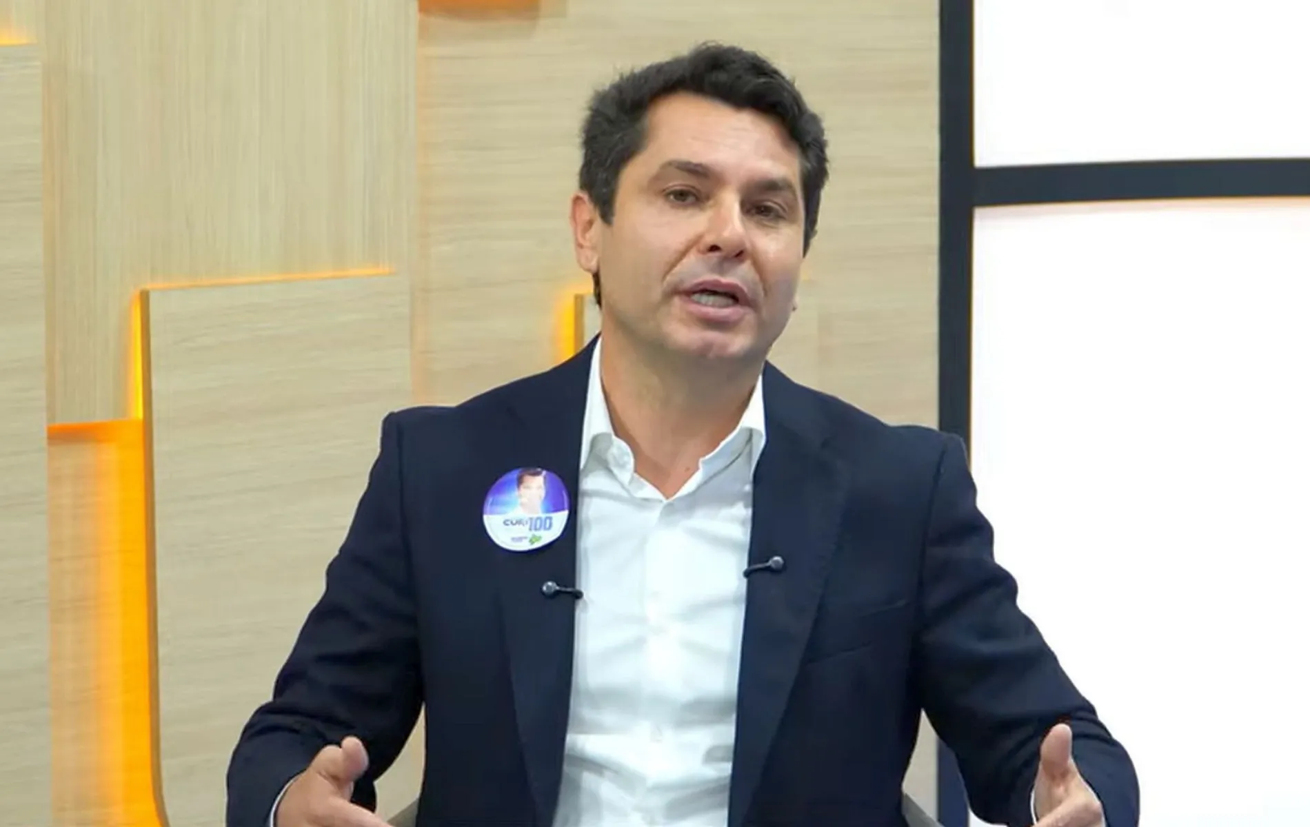 Fotografia em plano médio de um homem de pele clara, cabelos curtos castanho-escuros e olhos castanhos, sentado em um estúdio de televisão durante uma entrevista. Ele veste paletó escuro e camisa social branca de colarinho aberto. Na lapela direita do paletó, há um adesivo redondo de campanha eleitoral com sua foto e número. Ele está com as mãos gesticulando suavemente para a frente enquanto fala. Ao fundo, vê-se um painel de madeira com detalhes luminosos em tom alaranjado e um elemento gráfico branco à direita.