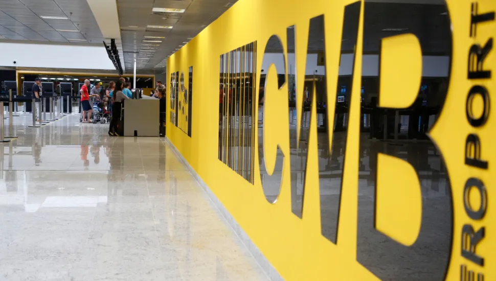 Fotografia em perspectiva de um saguão de aeroporto limpo e bem iluminado. Em primeiro plano, ao lado direito, destaca-se uma grande parede amarela com letras pretas em relevo onde se lê o código "CWB" seguido da palavra "AEROPORTO" disposta na vertical. Ao fundo, à esquerda, o piso claro e espelhado reflete as luzes do teto e a movimentação de passageiros, balcões de atendimento e filas de embarque.