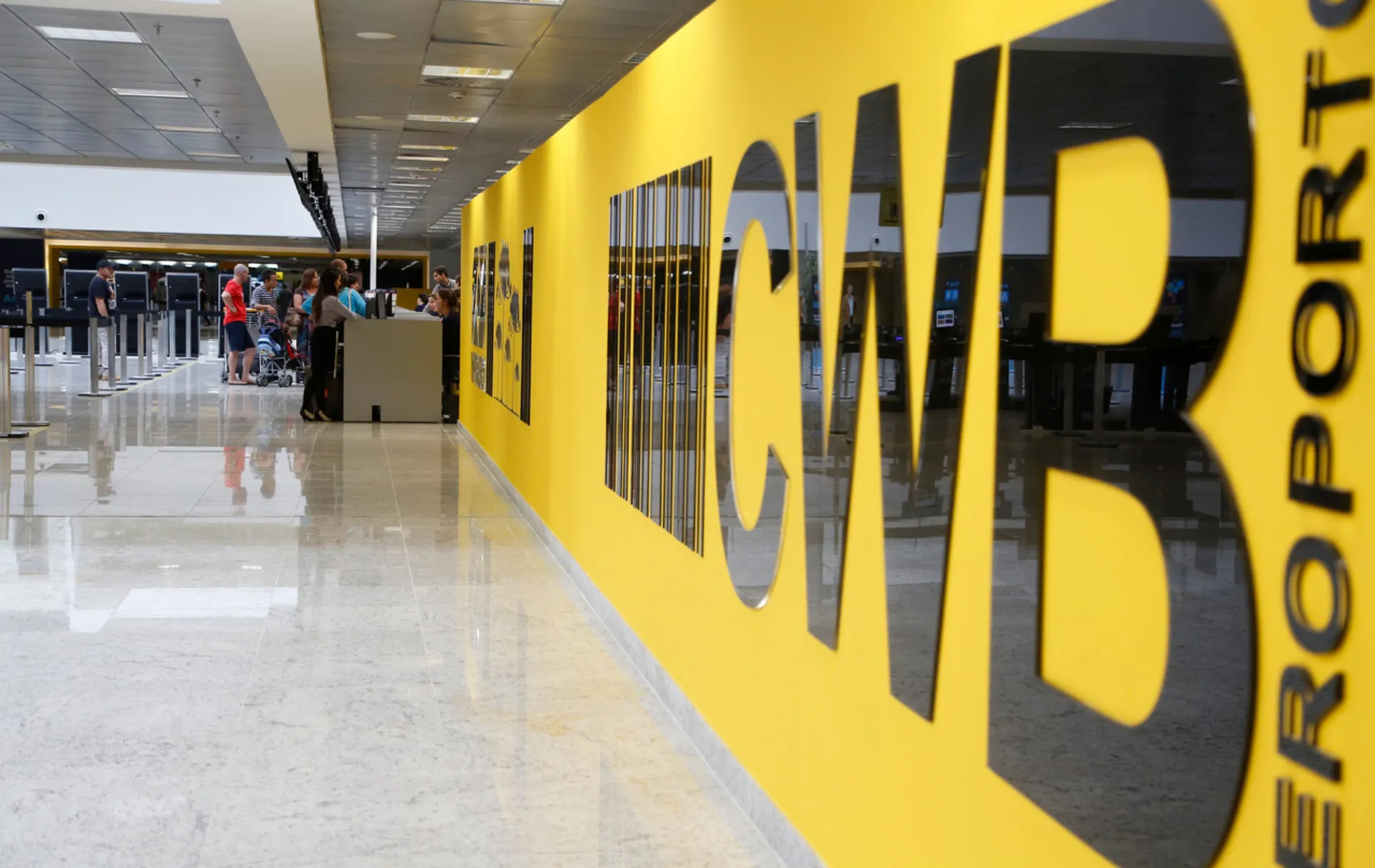 Fotografia em perspectiva de um saguão de aeroporto limpo e bem iluminado. Em primeiro plano, ao lado direito, destaca-se uma grande parede amarela com letras pretas em relevo onde se lê o código "CWB" seguido da palavra "AEROPORTO" disposta na vertical. Ao fundo, à esquerda, o piso claro e espelhado reflete as luzes do teto e a movimentação de passageiros, balcões de atendimento e filas de embarque.