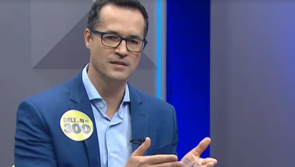 Fotografia em plano médio de Deltan Dallagnol, um homem de pele clara, cabelos curtos escuros e óculos de armação preta, sentado em um estúdio de televisão. Ele veste terno azul-petróleo e camisa social azul-clara sem gravata. Na lapela direita do terno, há um adesivo circular amarelo com o nome "DELTAN" e o número "300". Ele está com as mãos abertas gesticulando enquanto fala, mostrando anotações escritas a caneta na palma de sua mão esquerda. O fundo do estúdio é composto por painéis neutros em tons de cinza e azul.