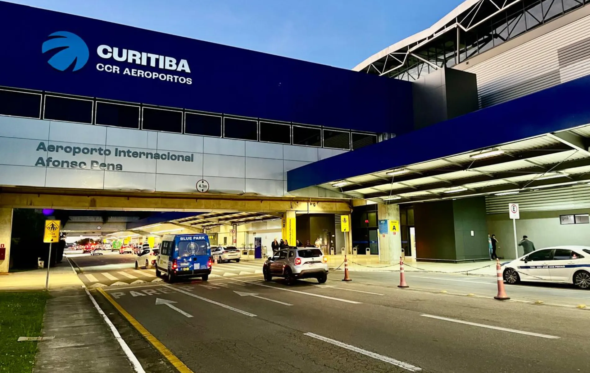 Fotografia em enquadramento aberto da fachada externa e do nível de embarque do Aeroporto Internacional Afonso Pena. No topo da estrutura moderna, destaca-se um grande painel azul com o logotipo e o nome "CURITIBA - CCR AEROPORTOS" em letras brancas, e logo abaixo a inscrição "Aeroporto Internacional Afonso Pena". Em primeiro plano, vê-se a via de acesso de veículos com sinalização horizontal no asfalto e cones de trânsito alaranjados delimitando faixas. Diversos carros e uma van azul estão parados ao longo do meio-fio para embarque e desembarque de passageiros.