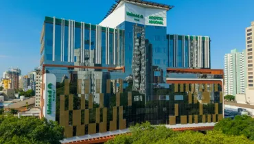 Fotografia em enquadramento amplo do prédio do Hospital Regional Unimed Maringá. A arquitetura moderna possui estrutura revestida por vidros espelhados azuis, detalhes em madeira e brises metálicos verticais e dourados. Na fachada superior e na lateral esquerda, sobressaem-se os logotipos verdes da "Unimed Maringá" e a inscrição "HOSPITAL REGIONAL". O edifício surge em meio à vegetação densa de árvores em primeiro plano, com outros prédios do centro urbano visíveis ao fundo sob céu azul limpo.