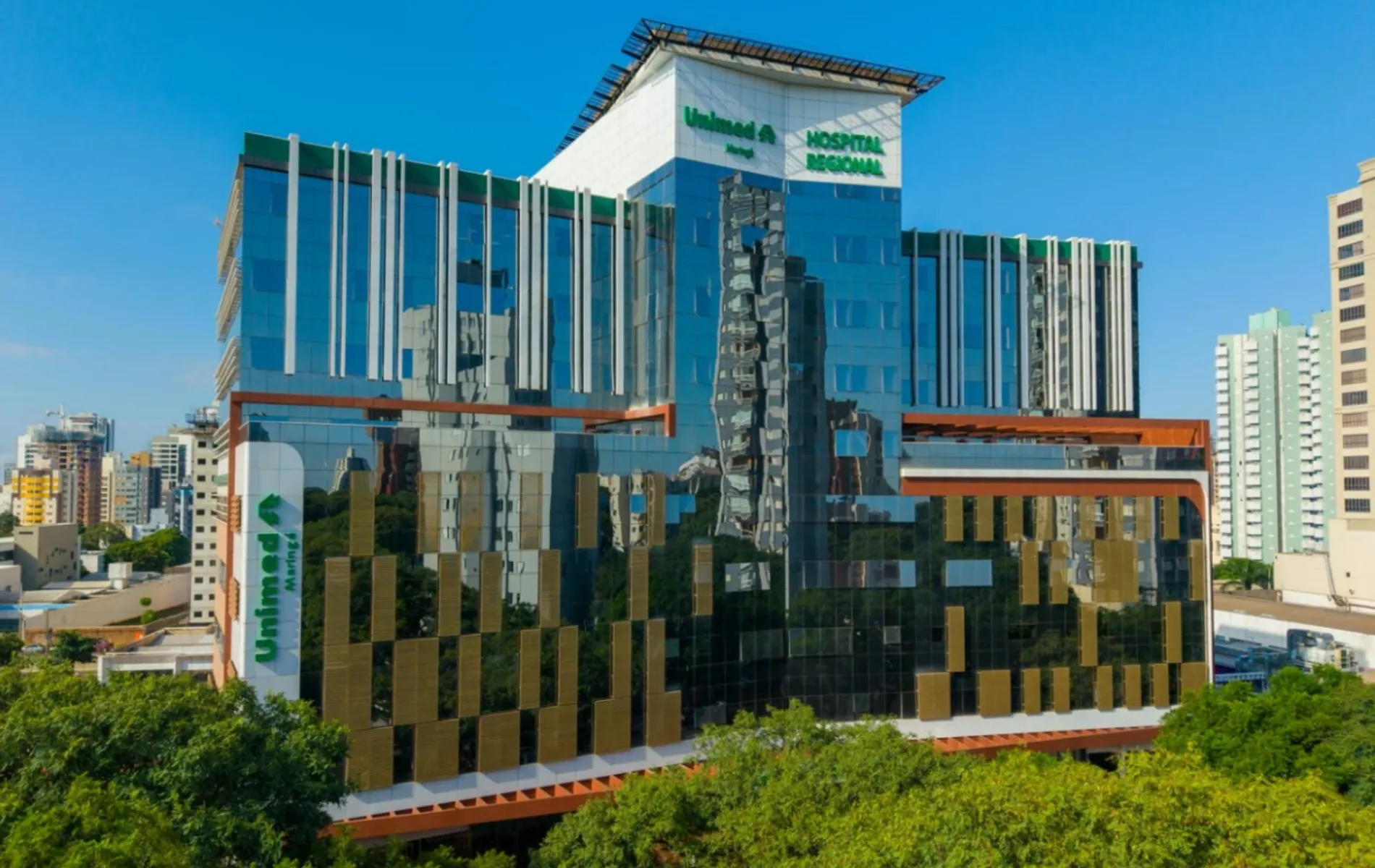 Fotografia em enquadramento amplo do prédio do Hospital Regional Unimed Maringá. A arquitetura moderna possui estrutura revestida por vidros espelhados azuis, detalhes em madeira e brises metálicos verticais e dourados. Na fachada superior e na lateral esquerda, sobressaem-se os logotipos verdes da "Unimed Maringá" e a inscrição "HOSPITAL REGIONAL". O edifício surge em meio à vegetação densa de árvores em primeiro plano, com outros prédios do centro urbano visíveis ao fundo sob céu azul limpo.