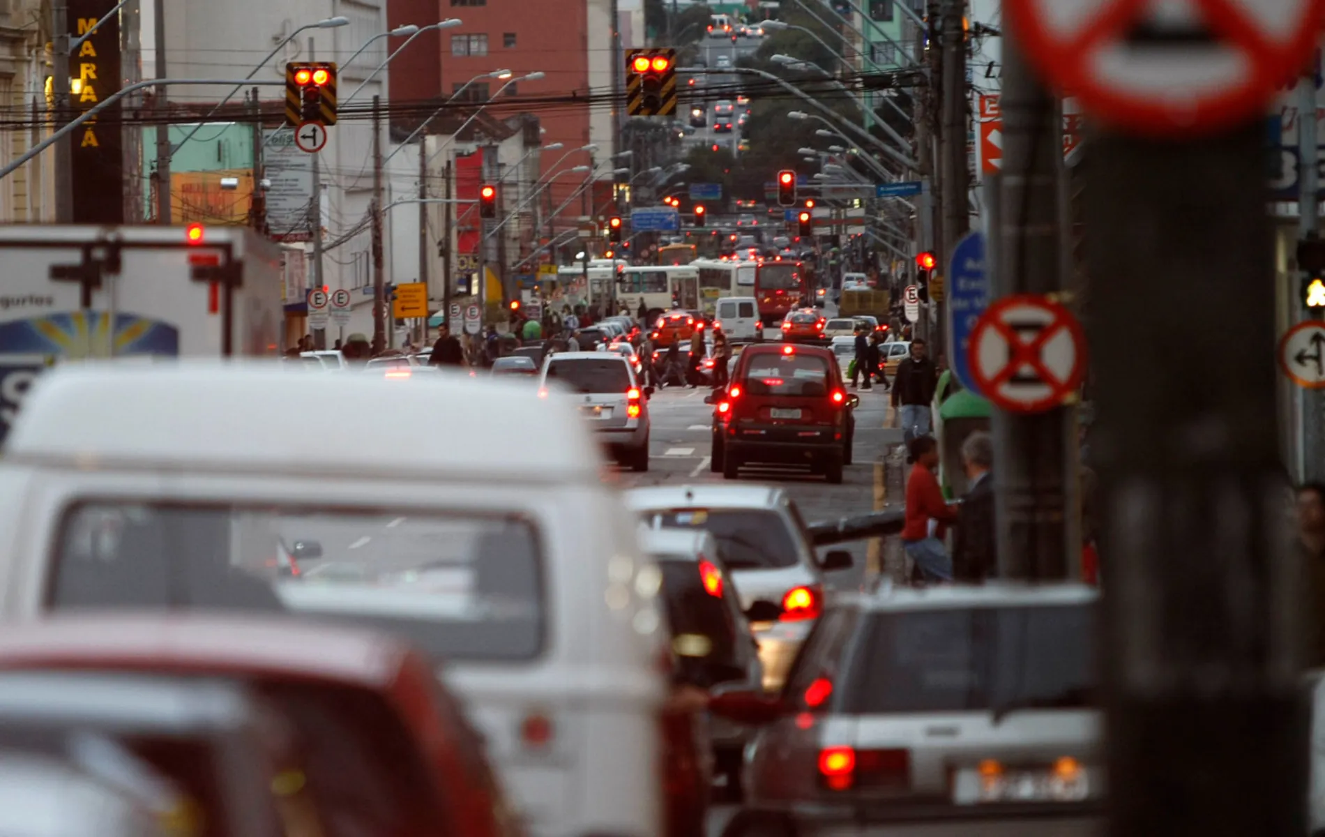 Fotografia em plano profundo capturada por lente teleobjetiva de uma movimentada avenida urbana durante o trânsito do fim da tarde. Em primeiro plano e ao longo da via, diversos veículos como carros e uma perua branca aparecem emparelhados, com luzes de freio e faróis acesos. Ao fundo, ao longo do relevo inclinado da rua, alinham-se múltiplos semáforos iluminados em vermelho, fiação elétrica, placas de trânsito e pedestres nas calçadas sob iluminação natural suave.