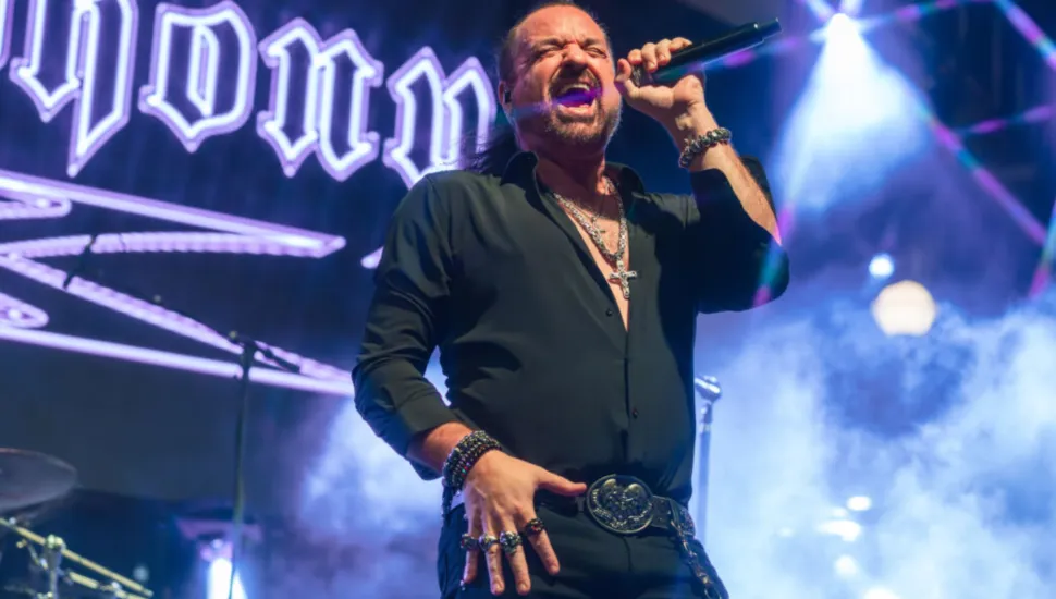 Fotografia em enquadramento médio de um cantor de rock no palco durante uma apresentação ao vivo. O homem, de pele clara, cabelos escuros presos para trás e barba cavanhaque, está com a boca aberta cantando em um microfone que segura na mão esquerda. Ele veste camisa preta de mangas compridas dobradas até os antebraços, aberta no peito exibindo correntes e crucifixos prateados, calça escura e cinto com fivela grande decorada. Em sua mão direita, apoia a palma sobre a coxa, exibindo anéis e pulseiras. Ao fundo, há holofotes acesos, luzes roxas e azuladas e fumaça cênica sob o letreiro do show.