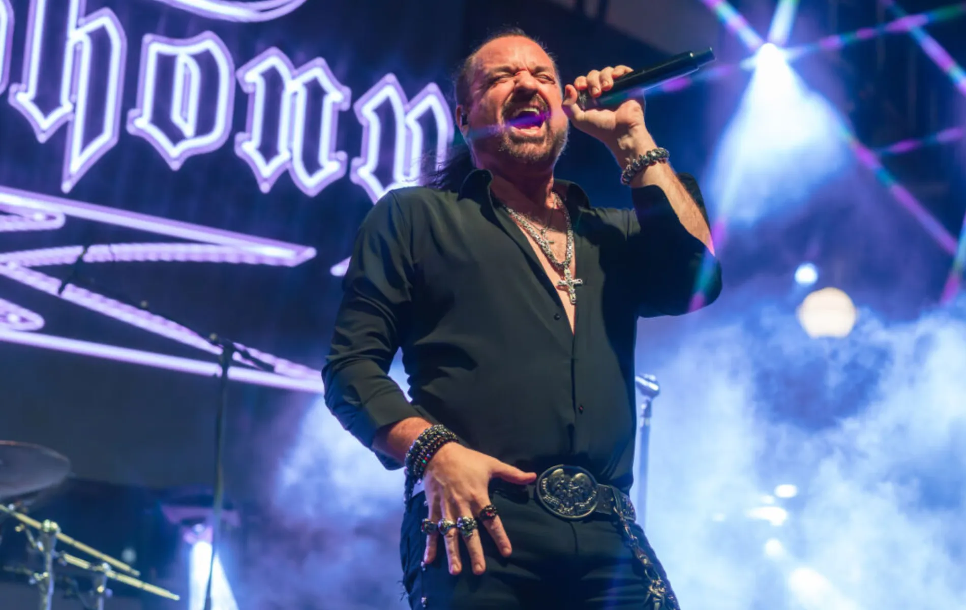 Fotografia em enquadramento médio de um cantor de rock no palco durante uma apresentação ao vivo. O homem, de pele clara, cabelos escuros presos para trás e barba cavanhaque, está com a boca aberta cantando em um microfone que segura na mão esquerda. Ele veste camisa preta de mangas compridas dobradas até os antebraços, aberta no peito exibindo correntes e crucifixos prateados, calça escura e cinto com fivela grande decorada. Em sua mão direita, apoia a palma sobre a coxa, exibindo anéis e pulseiras. Ao fundo, há holofotes acesos, luzes roxas e azuladas e fumaça cênica sob o letreiro do show.