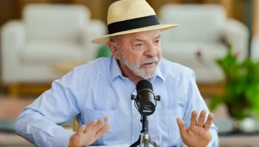Fotografia em plano médio do presidente Luiz Inácio Lula da Silva, um homem idoso de pele clara, cabelos e barba grisalhos. Ele veste uma camisa guayabera azul-clara de mangas compridas e um chapéu panamá bege com fita preta. Lula está sentado à mesa, com as mãos espalmadas e abertas para a frente enquanto fala em um microfone profissional preto montado em um pequeno tripé à sua frente. Ao fundo, desfocado, vê-se um ambiente residencial com poltronas claras e plantas decorativas.