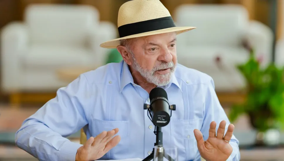 Fotografia em plano médio do presidente Luiz Inácio Lula da Silva, um homem idoso de pele clara, cabelos e barba grisalhos. Ele veste uma camisa guayabera azul-clara de mangas compridas e um chapéu panamá bege com fita preta. Lula está sentado à mesa, com as mãos espalmadas e abertas para a frente enquanto fala em um microfone profissional preto montado em um pequeno tripé à sua frente. Ao fundo, desfocado, vê-se um ambiente residencial com poltronas claras e plantas decorativas.