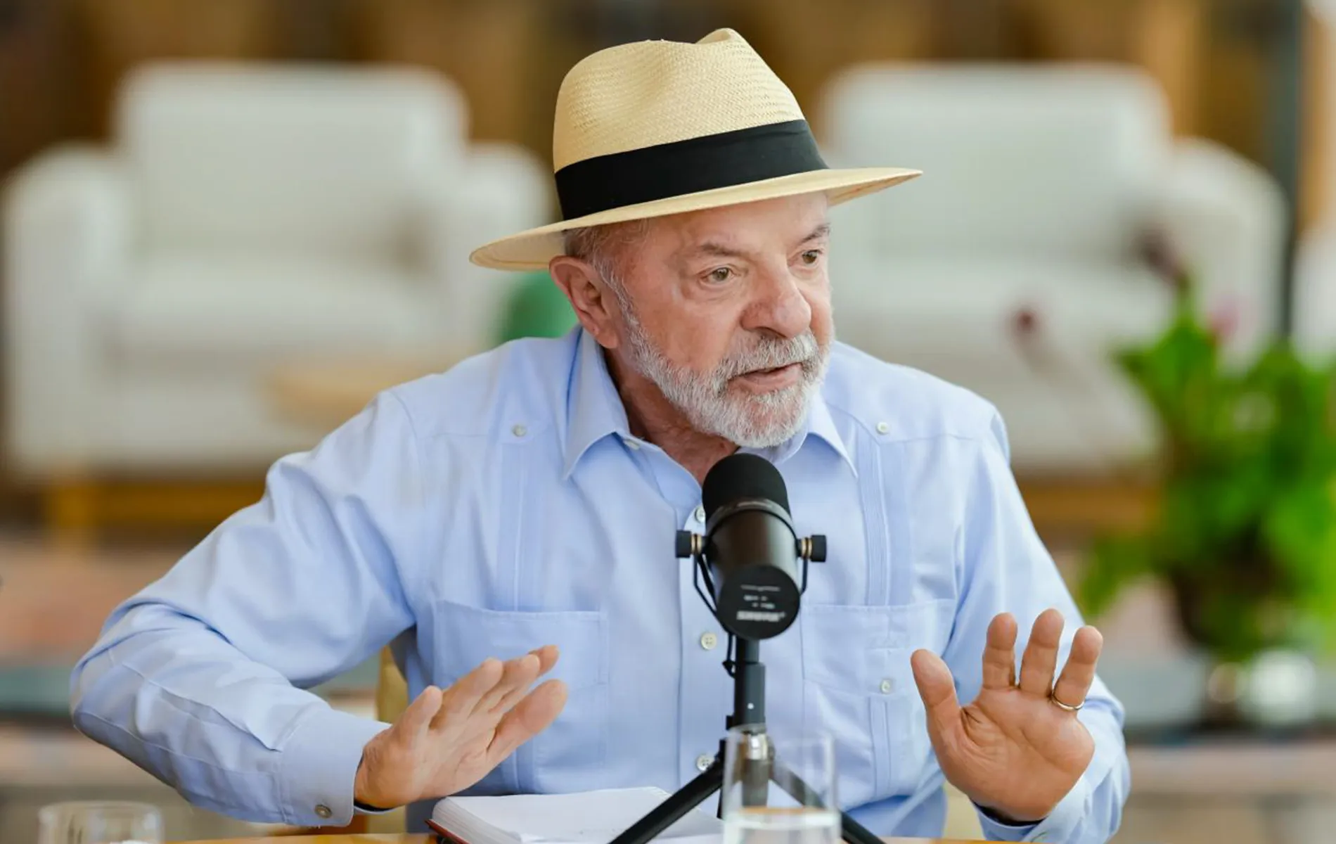 Fotografia em plano médio do presidente Luiz Inácio Lula da Silva, um homem idoso de pele clara, cabelos e barba grisalhos. Ele veste uma camisa guayabera azul-clara de mangas compridas e um chapéu panamá bege com fita preta. Lula está sentado à mesa, com as mãos espalmadas e abertas para a frente enquanto fala em um microfone profissional preto montado em um pequeno tripé à sua frente. Ao fundo, desfocado, vê-se um ambiente residencial com poltronas claras e plantas decorativas.