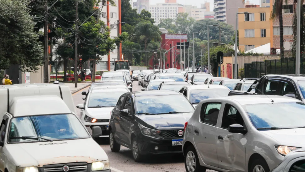 Fotografia em enquadramento frontal e plano fechado de um congestionamento de trânsito em uma rua urbana. Diversos automóveis em cores neutras — como prata, cinza, branco e preto — estão enfileirados lado a lado, ocupando totalmente as faixas de rodagem. Em primeiro plano, destacam-se uma furgoneta branca à esquerda, um hatchback cinza-escuro ao centro e um carro compacto prata à direita. Ao fundo, veem-se calçadas arborizadas, fiação aérea e edifícios residenciais sob luz natural difusa de dia nublado.