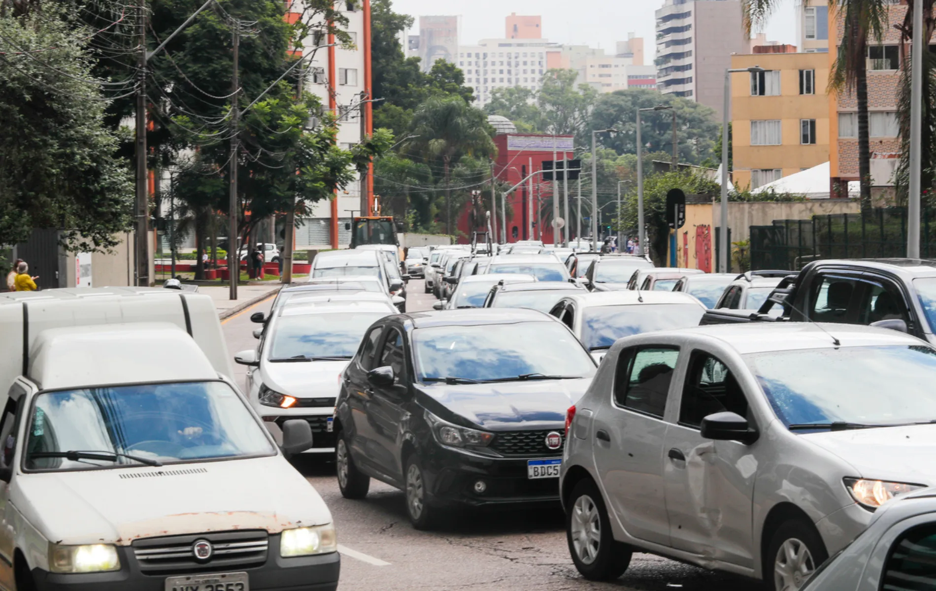 Fotografia em enquadramento frontal e plano fechado de um congestionamento de trânsito em uma rua urbana. Diversos automóveis em cores neutras — como prata, cinza, branco e preto — estão enfileirados lado a lado, ocupando totalmente as faixas de rodagem. Em primeiro plano, destacam-se uma furgoneta branca à esquerda, um hatchback cinza-escuro ao centro e um carro compacto prata à direita. Ao fundo, veem-se calçadas arborizadas, fiação aérea e edifícios residenciais sob luz natural difusa de dia nublado.
