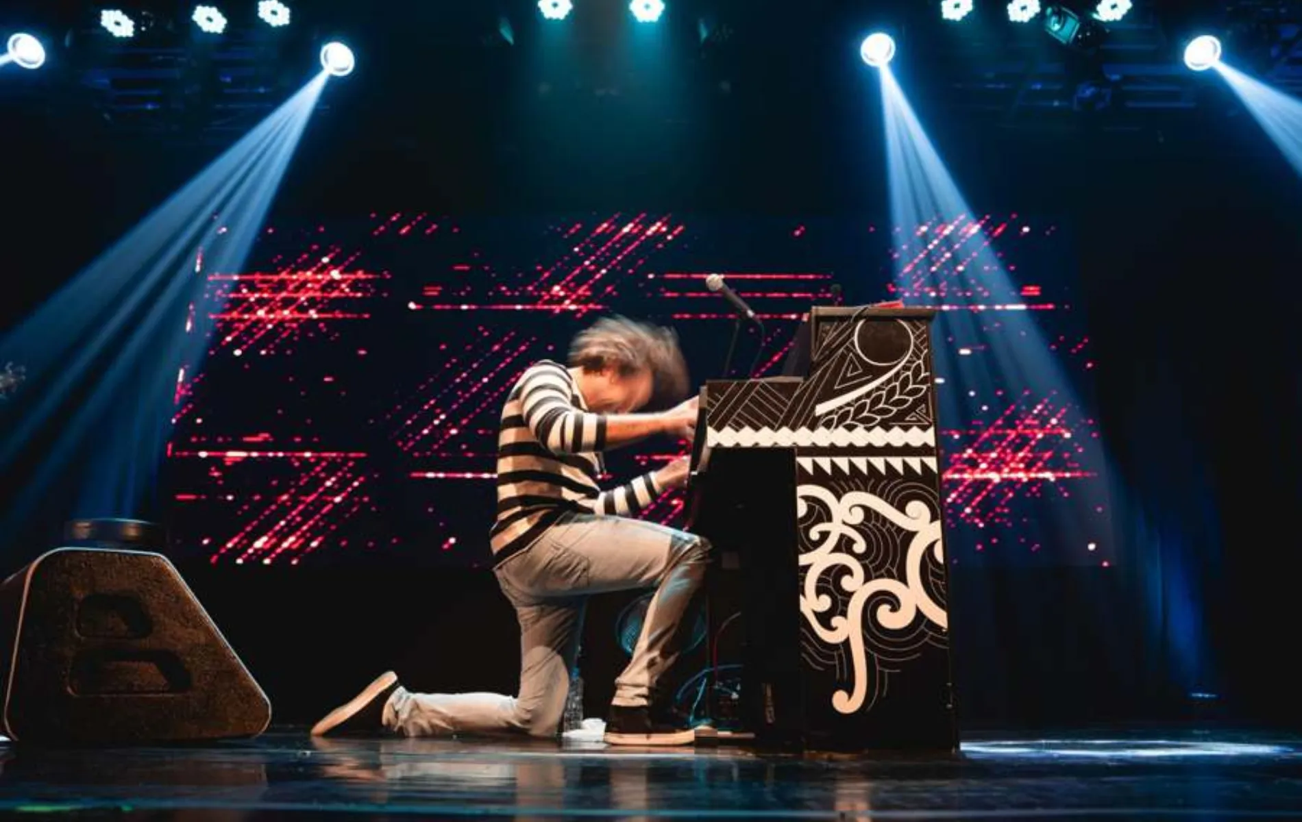 Fotografia em plano geral e ângulo baixo de um músico se apresentando no palco durante um show. O homem está ajoelhado no chão de joelhos dobrados, inclinado para a frente em movimento vigoroso enquanto toca um piano vertical artisticamente decorado com grafismos pretos e brancos. Ele veste blusa de mangas compridas listrada em preto e branco, calça jeans e tênis. Ao fundo, um painel de LED exibe luzes vermelhas em padrões abstratos, contrastando com os feixes de luz azul-clara vindos dos holofotes no teto. À esquerda do palco, sobressai-se uma caixa de retorno de som preta.