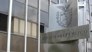 Fotografia em enquadramento aproximado e ângulo em contra-plongée da entrada de um edifício público. Em primeiro plano, à direita, destaca-se uma placa metálica escovada na cor prata onde está impresso o brasão de armas do estado e a inscrição "MINISTÉRIO PÚBLICO do Estado do Paraná". Ao fundo, à esquerda, vê-se a fachada do prédio composta por grandes janelas de vidro espelhado dispostas em uma estrutura com vigas metálicas verticais e prateadas.