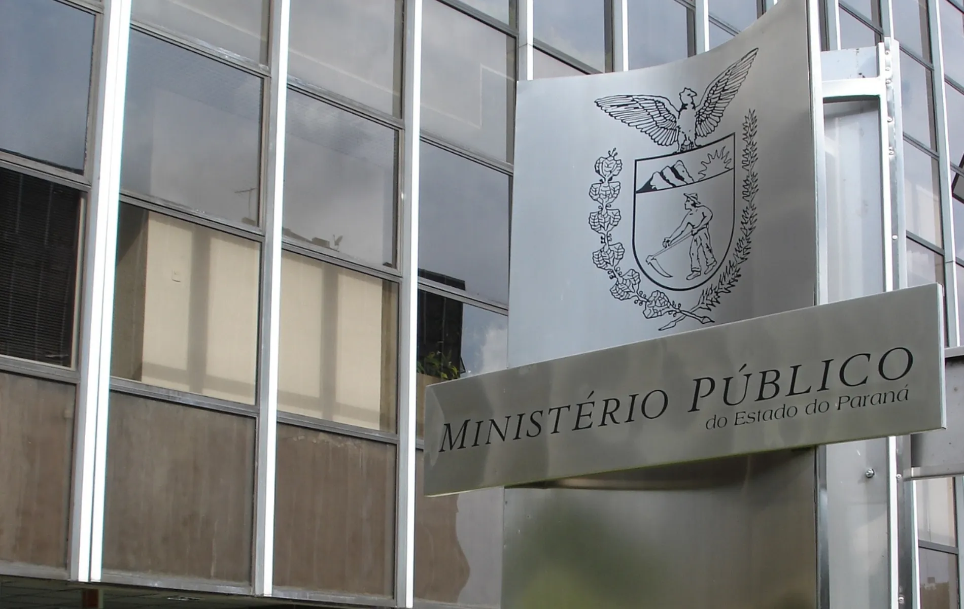 Fotografia em enquadramento aproximado e ângulo em contra-plongée da entrada de um edifício público. Em primeiro plano, à direita, destaca-se uma placa metálica escovada na cor prata onde está impresso o brasão de armas do estado e a inscrição "MINISTÉRIO PÚBLICO do Estado do Paraná". Ao fundo, à esquerda, vê-se a fachada do prédio composta por grandes janelas de vidro espelhado dispostas em uma estrutura com vigas metálicas verticais e prateadas.