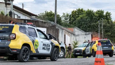 Fotografia em enquadramento médio e ângulo baixo de viaturas da Polícia Militar do Paraná estacionadas em uma rua residencial. Em primeiro plano, à esquerda, destaca-se a lateral de uma SUV amarela, cinza e branca com a inscrição "POLÍCIA MILITAR 10º BPM" e o brasão do estado na porta. Em segundo plano, no centro, um policial militar fardado e de boina preta está de pé ao lado de outro veículo policial. Ao fundo, à direita, outra viatura com os giroflex acesos em vermelho estaciona próximo a um muro alto com arame de proteção. Em primeiro plano, à extrema direita, há um cone de trânsito alaranjado com a sigla "PMPR".